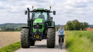 Eine Dame fährt neben einem großen Traktor Rad. Die Dame trägt ein hellblaues T-Shirt, einen Helm. Sie ist blond und etwa 35 Jahre alt.