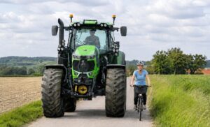 Auf dem Bild ist ein großer grüner Traktor der Marke Deutz Vario zu sehen. Rechts davon fährt eine Radfahrerin, blond, attraktiv, ca. 35 Jahre alt. Sie trägt ein hellblaues Shirt und eine schwarze, lange Hose sowie einen grauen Fahrradhelm. Die Dame lächelt und sieht zufrieden aus. Der Traktorfahrer ist männlich und hinter der etwas spiegelnden Scheibe nur schemenhaft zu erkennen. Die Szene befindet sich im ländlichen Raum, links ein abgeerntetes Getreidefeld, rechts Grünland. Das Wetter ist heiter bewölkt.