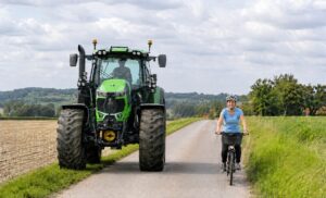 Auf dem Bild ist ein großer grüner Traktor der Marke Deutz Vario zu sehen. Rechts davon fährt eine Radfahrerin, blond, attraktiv, ca. 35 Jahre alt. Sie trägt ein hellblaues Shirt und eine schwarze, lange Hose sowie einen grauen Fahrradhelm. Die Dame lächelt und sieht zufrieden aus. Der Traktorfahrer ist männlich und hinter der etwas spiegelnden Scheibe nur schemenhaft zu erkennen. Die Szene befindet sich im ländlichen Raum, links ein abgeerntetes Getreidefeld, rechts Grünland. Das Wetter ist heiter bewölkt.